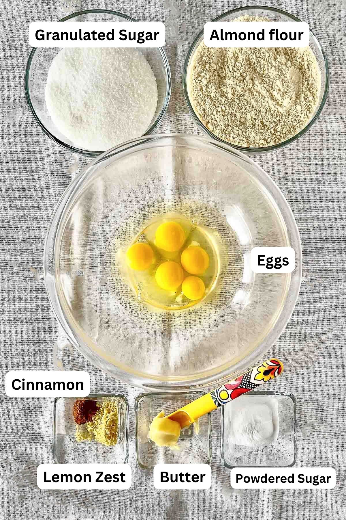 Ingredients on the table in bowls to make a spanish almond cake with text labels.