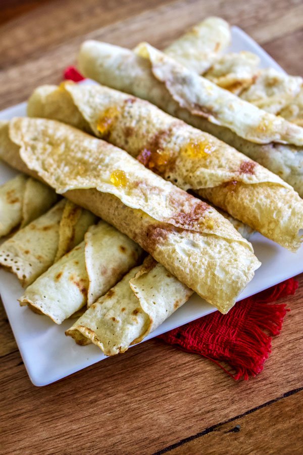 How To Make Crepes- The Easy Way- Rolled crepes filled with jam on a white plate.