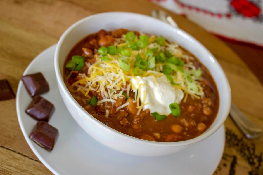 Never had chocolate chipotle chili soup before? Chili soup is a comfort food perfect for the cold nights. When you add smoky chipotle chiles and earthy unsweetened chocolate to it, you add unexpected depth and richness to this hearty chili. Chocolate Chipotle Chili Soup in a white bowl on a white plate with three pieces of chocolate and sour cream on top of soup