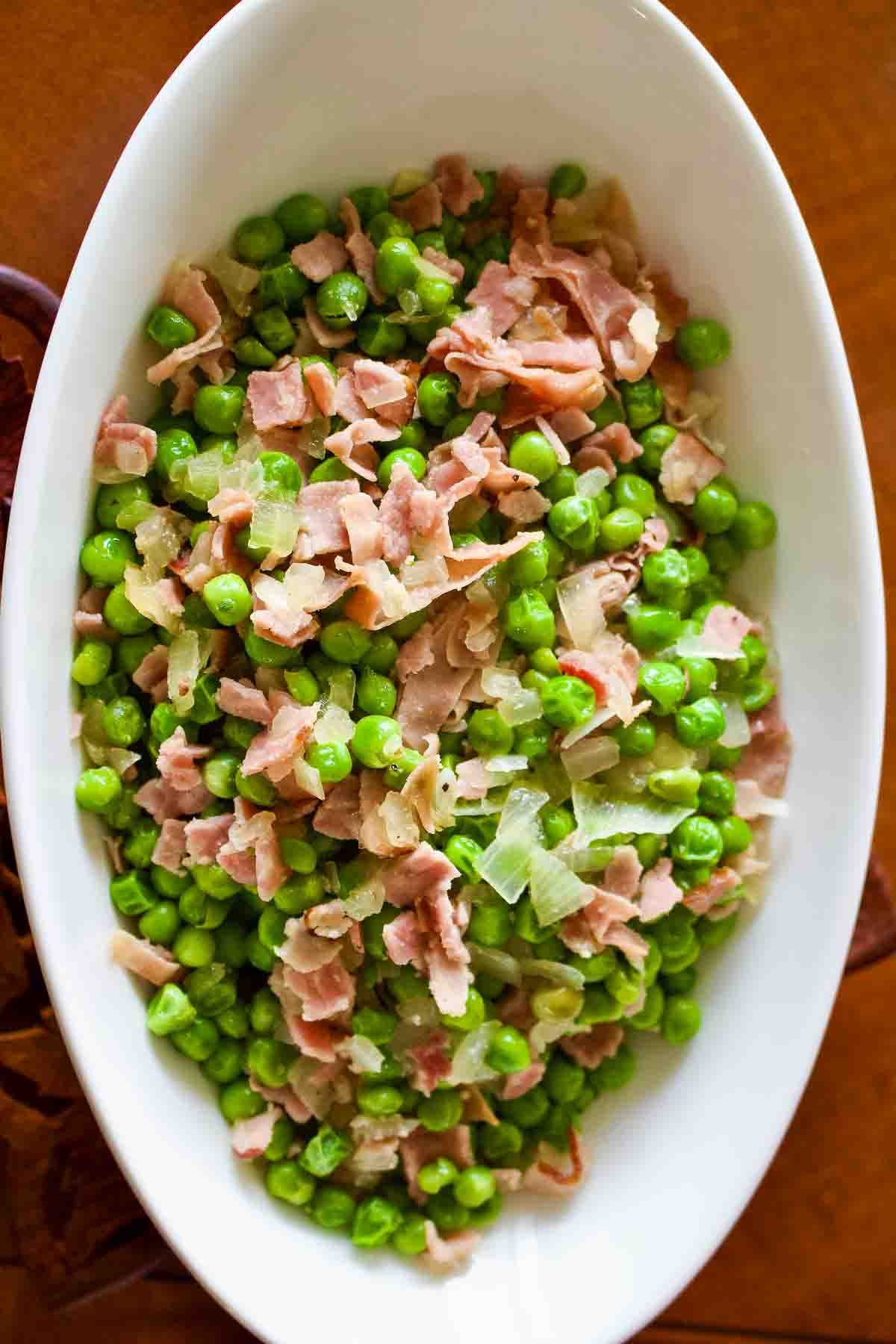 Easy Green Peas With Ham- Spanish recipe- is an easy dish that can be made in no time using fresh or frozen peas. For authenticity, use jamon Serrano. Peas With Ham in a white oval serving bowl.