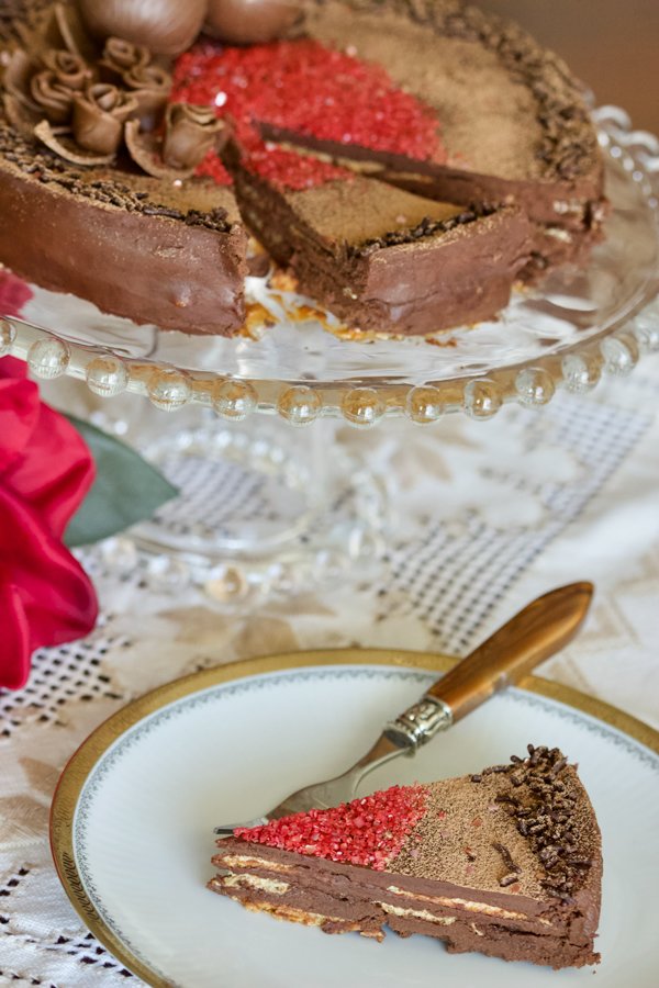 This is a classic European chocolate ganache meringue torte recipe. Easy to make but decadent, this recipe is the perfect option for the chocolate lovers. Classic European chocolate ganache meringue torte- slice of cake on a white plate with fork . Cake sitting on a cake platter in the background