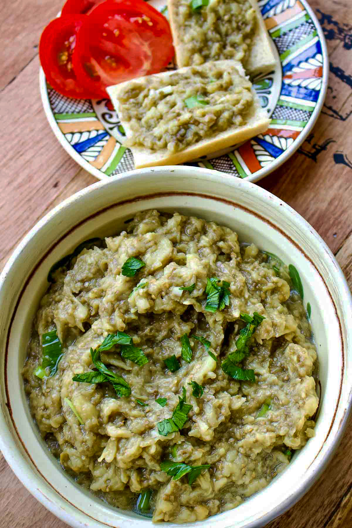 Roasted Eggplant Salad- Romanian Style is a delicious summer salad that can be served on fresh bread with tomatoes and sometimes Feta cheese. Roasted eggplant salad in a bowl and slices of bread with eggplant and tomato in the background.