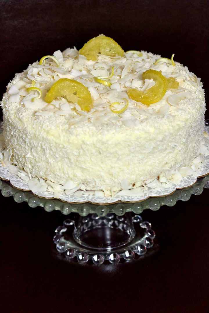 Cream cheese lemon coconut cake is a versatile recipe that can be made any time of the year. It is suitable for the holidays or any celebrations when you need a beautiful dessert that everyone can have. It has a subtle tropical taste from the lemon and the coconut combination. Lemon Cream Cheese Cake With Coconut sitting on a glass cake stand.
