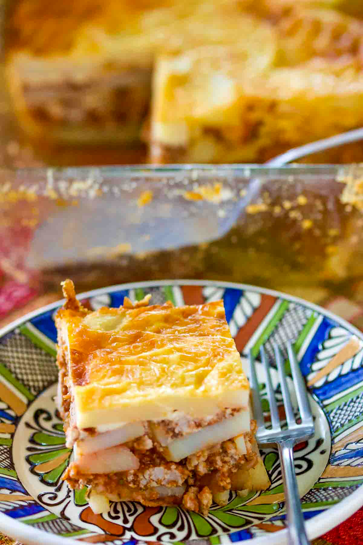 The Romanian Authentic Potato Moussaka is made with layers of potatoes and minced meat. This dish is comfort food, perfect for a family! Romanian Potato Moussaka on a traditional Romanian plate with fork.