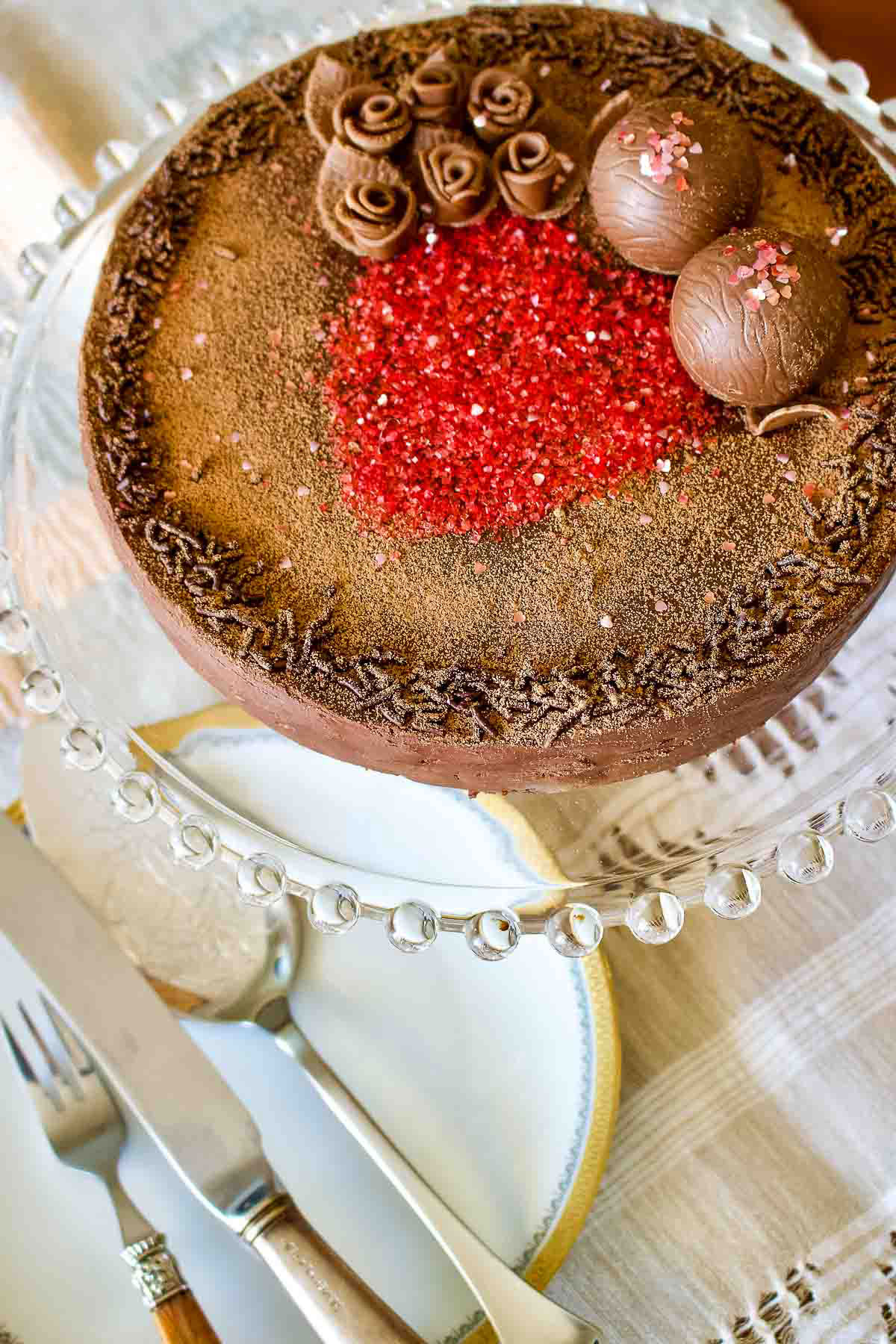 This is a classic European chocolate ganache meringue torte recipe. Easy to make but decadent, this recipe is the perfect option for the chocolate lovers. Decorated Meringue Cake With Chocolate Ganache sitting on a glass cake support.