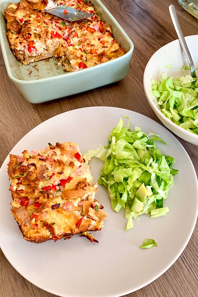 Plate with a slice of strata next to salad and strata baking dish in the background