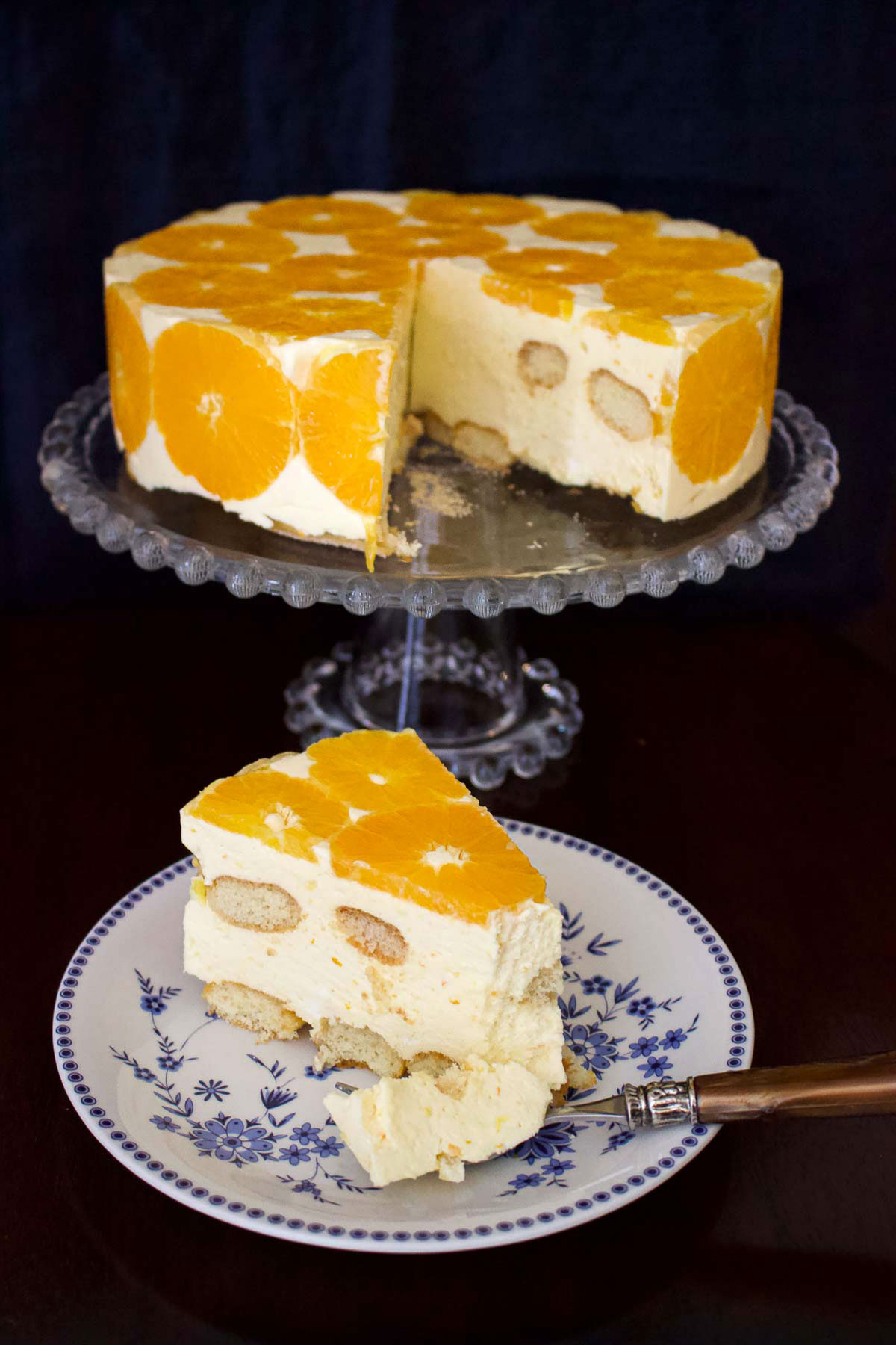 Orange Charlotte Cake slice on a blue and white plate with fork and cake on a glass cake stand0