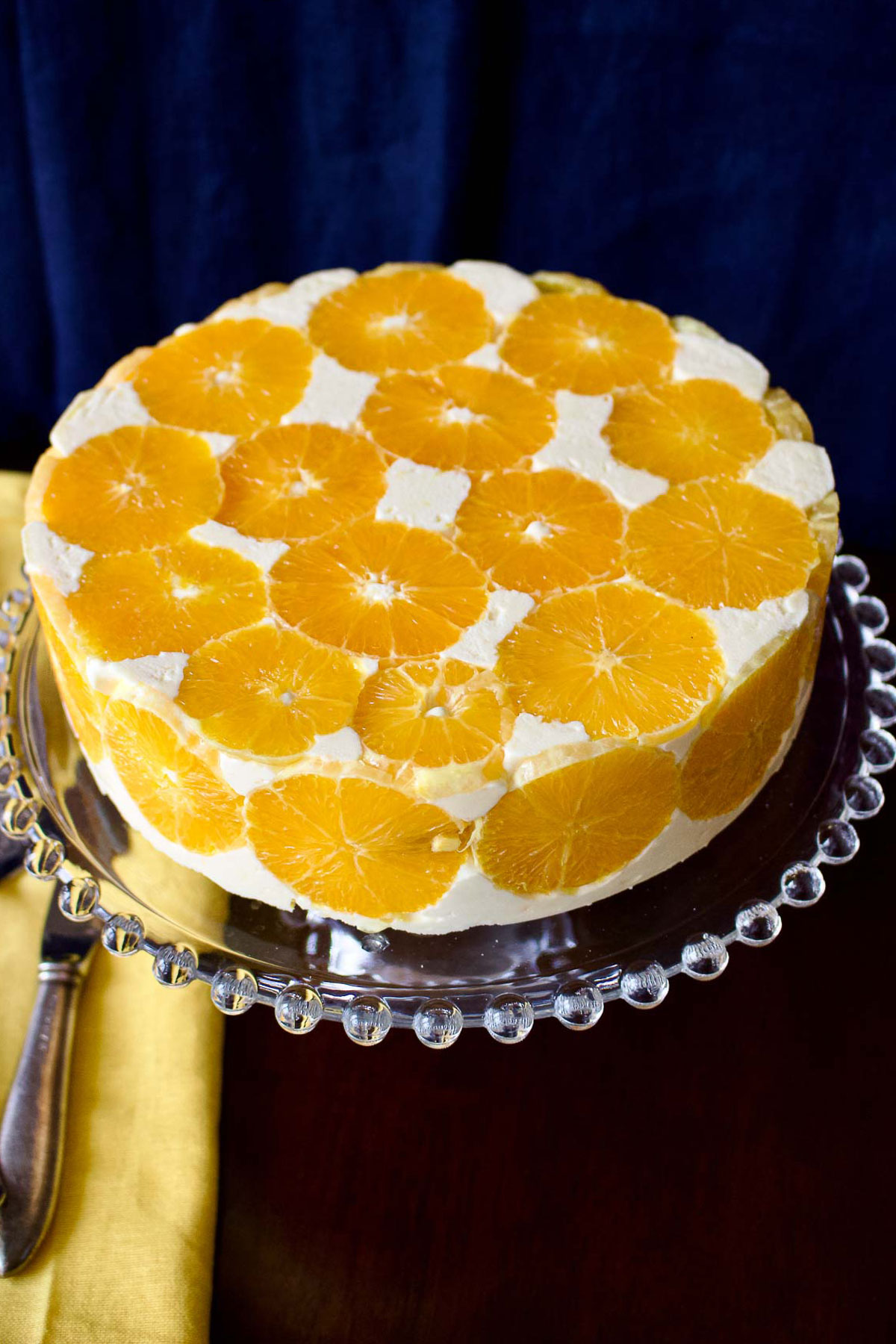 Delicious and beautiful decorated Orange Charlotte Cake sitting on a glass cake support with knife and yellow tea towel on the left.